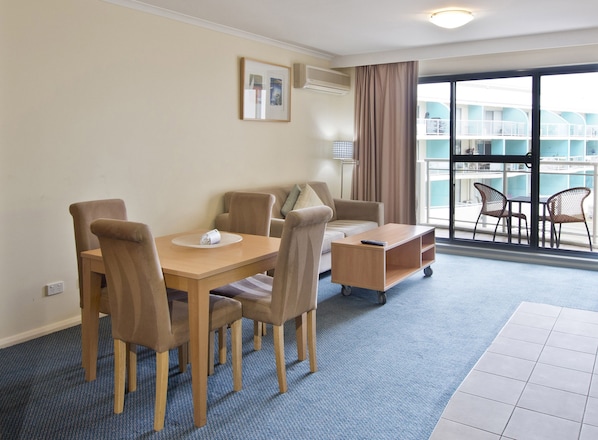 A hotel room with a dining table, chairs, a small sofa, a side table, and a view of a pool and building through large windows. - Oaks The Entrance Waterfront Suites (The Entrance)