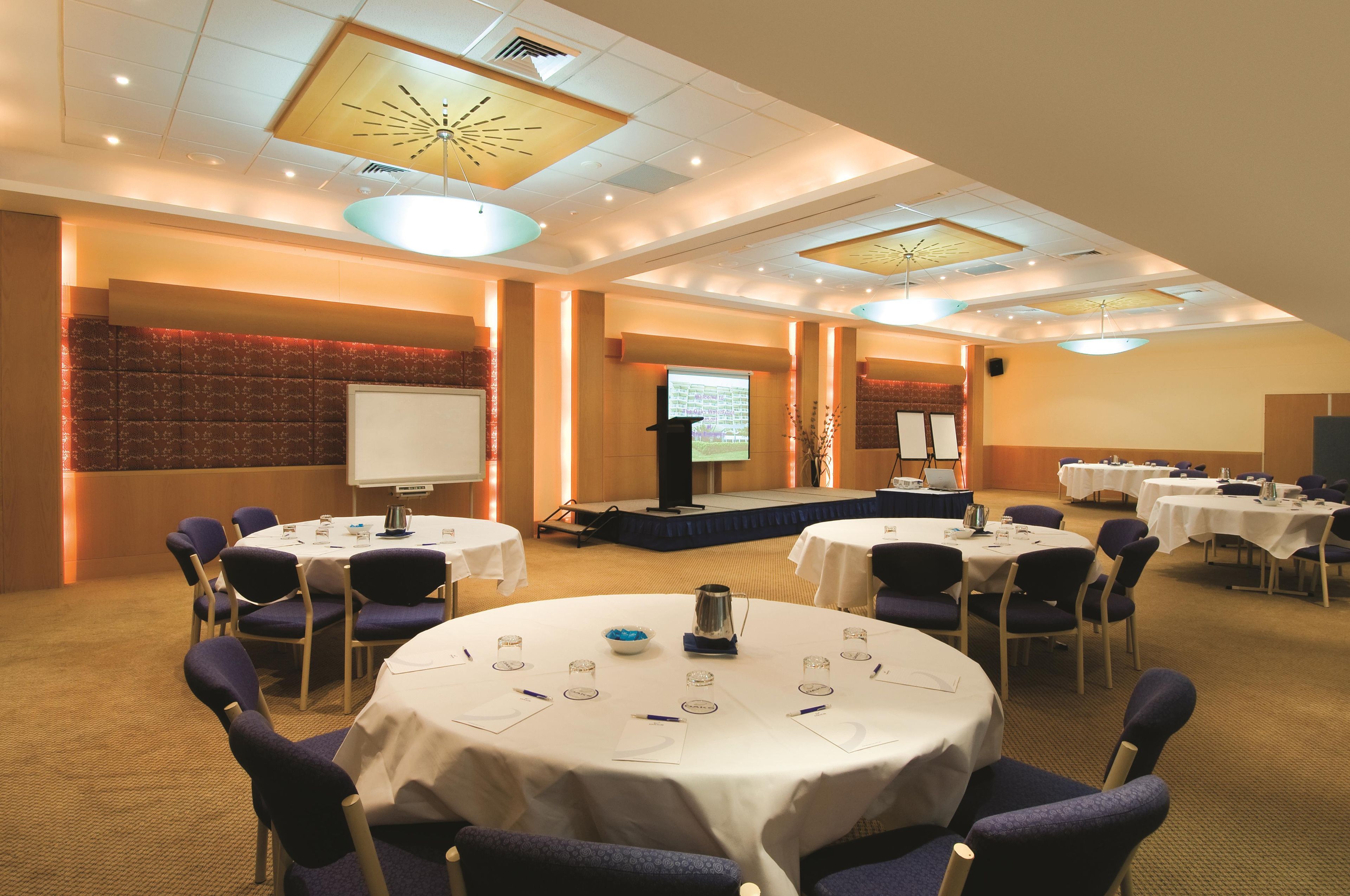 A conference room with multiple tables, chairs, a stage, and a presentation screen.