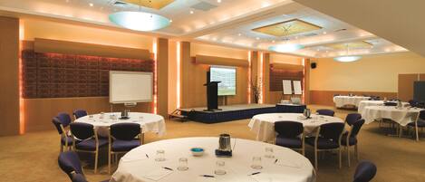 A conference room with multiple tables, chairs, a stage, and a presentation screen.