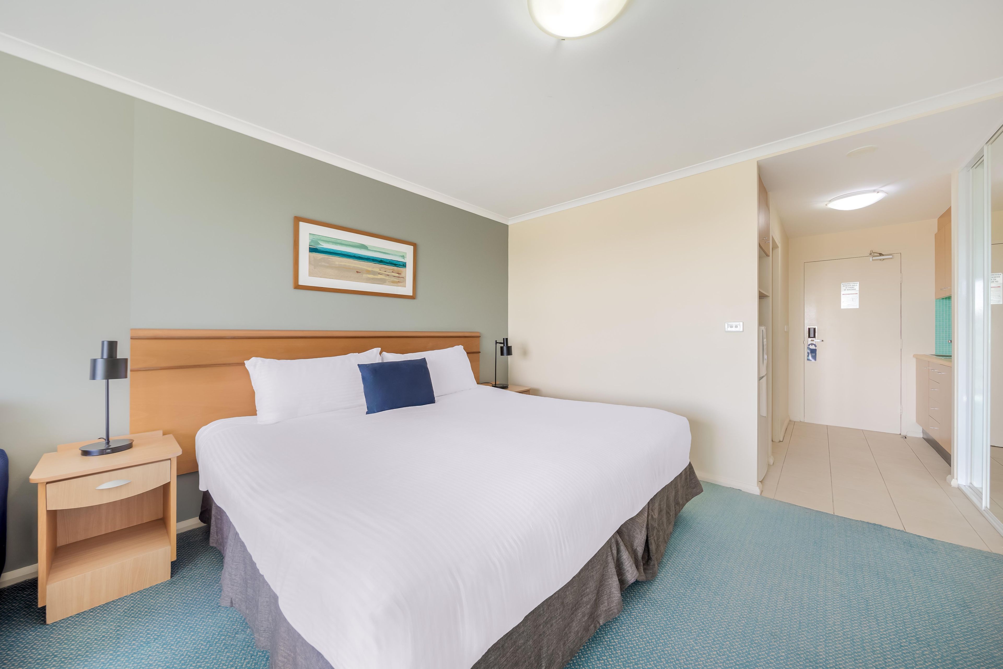 Habitación de hotel con una cama grande, una mesita de noche, una lámpara, un cuadro en la pared y un baño visible a través de una puerta abierta.