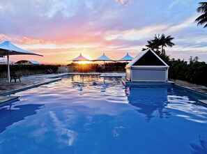 A swimming pool with a sunset in the background, surrounded by umbrellas and palm trees.