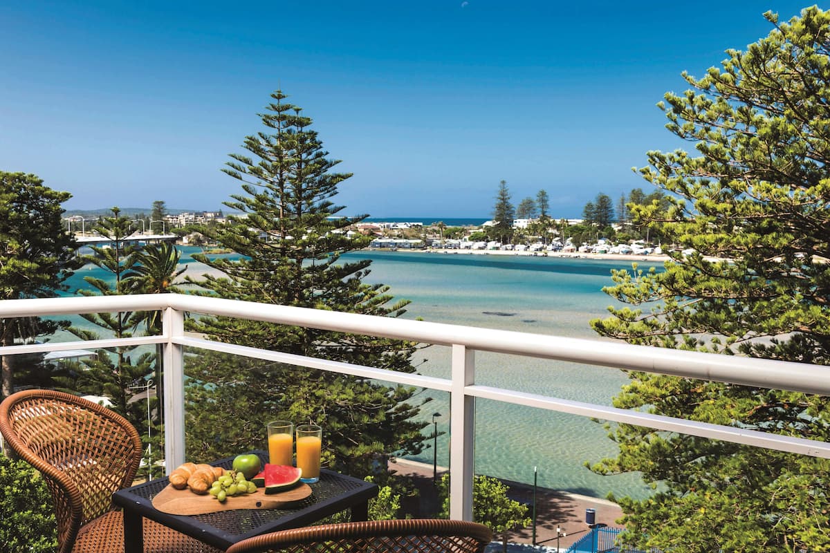 A balcony with a wicker chair, a wooden table with fruits and a glass of juice, overlooking a clear blue sea and a beach with trees and buildings in the background.