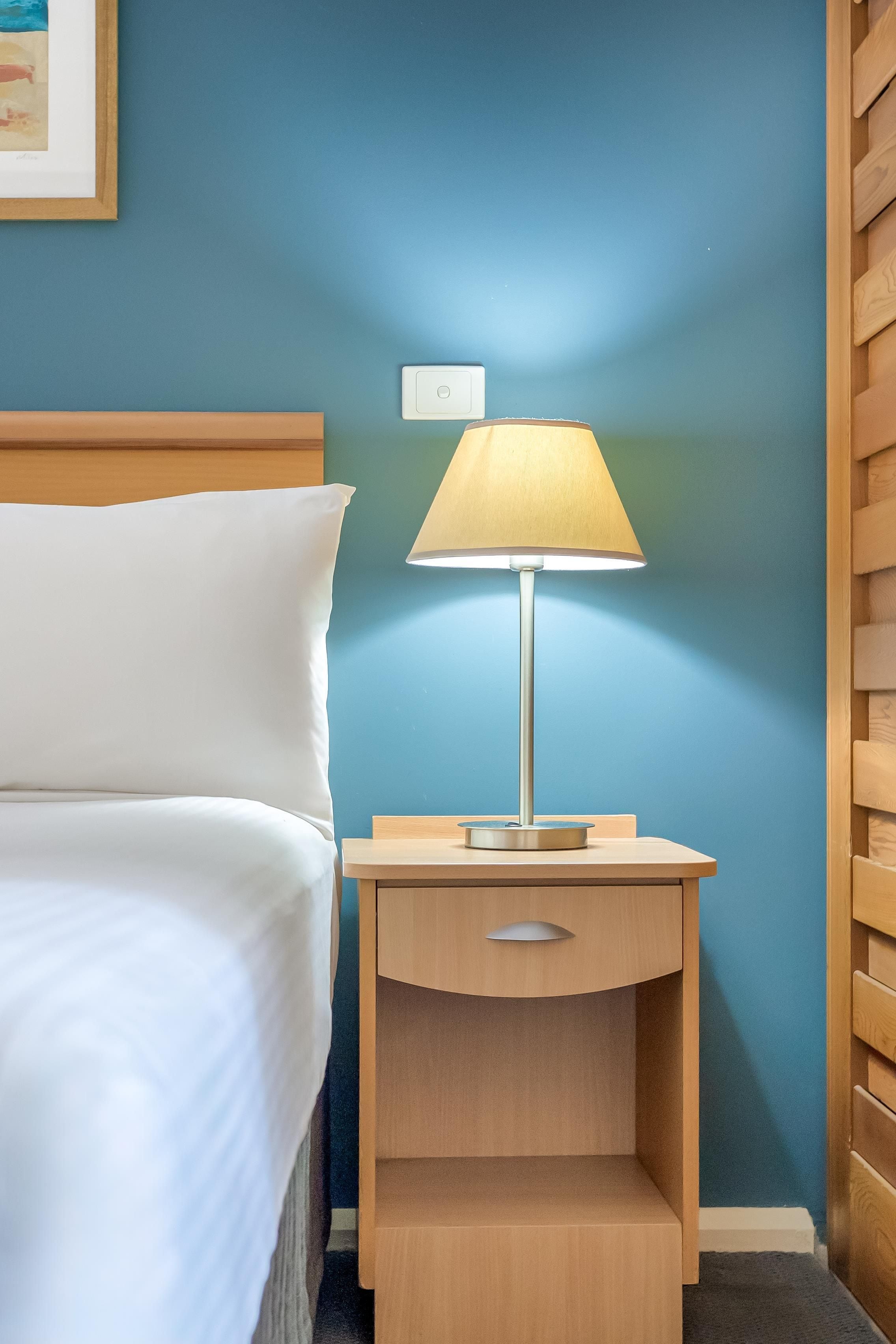 A neatly made bed with white linens, a wooden nightstand with a lamp, and a wall-mounted light switch.