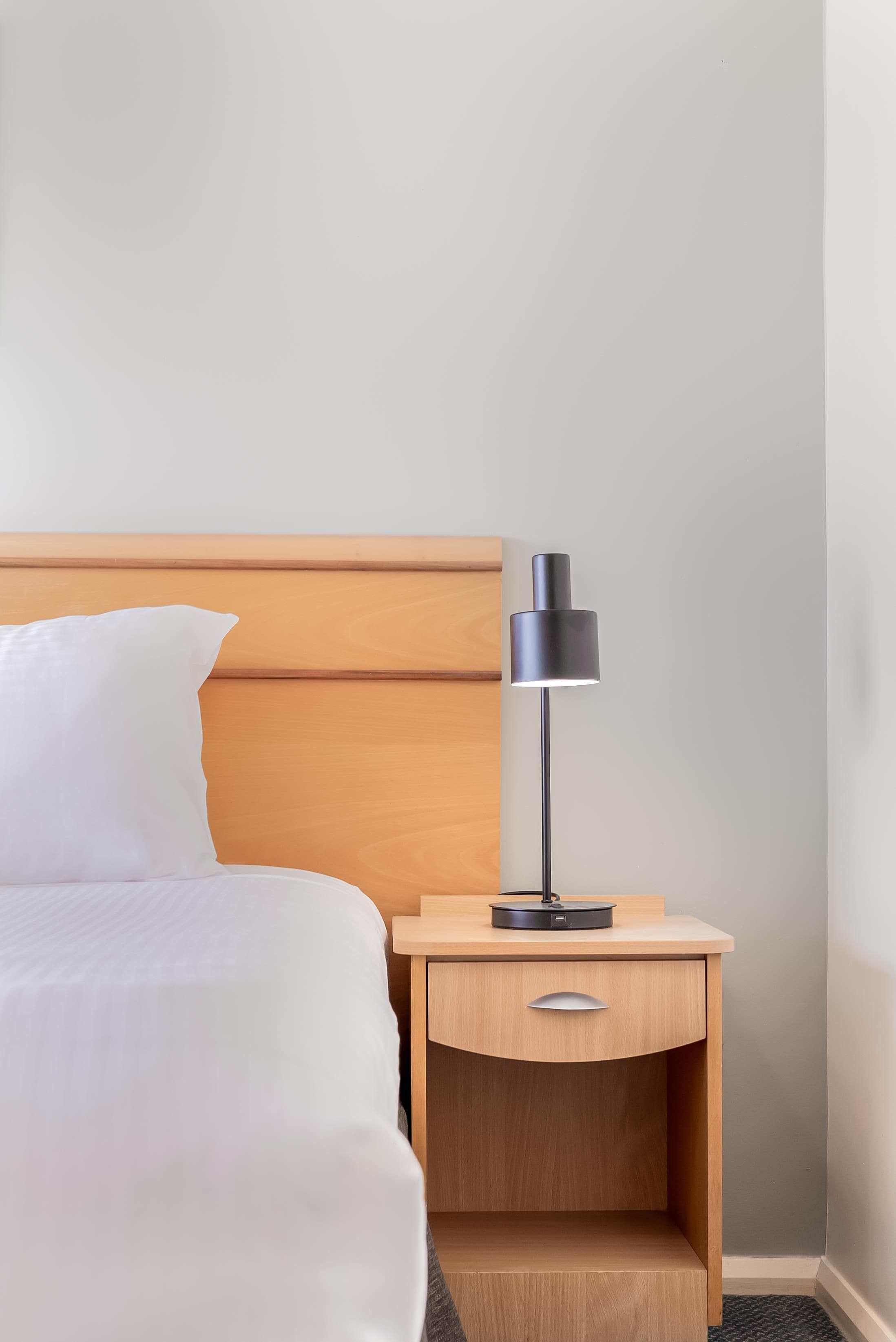A hotel room with a bed, a bedside table with a lamp, and a wooden headboard.