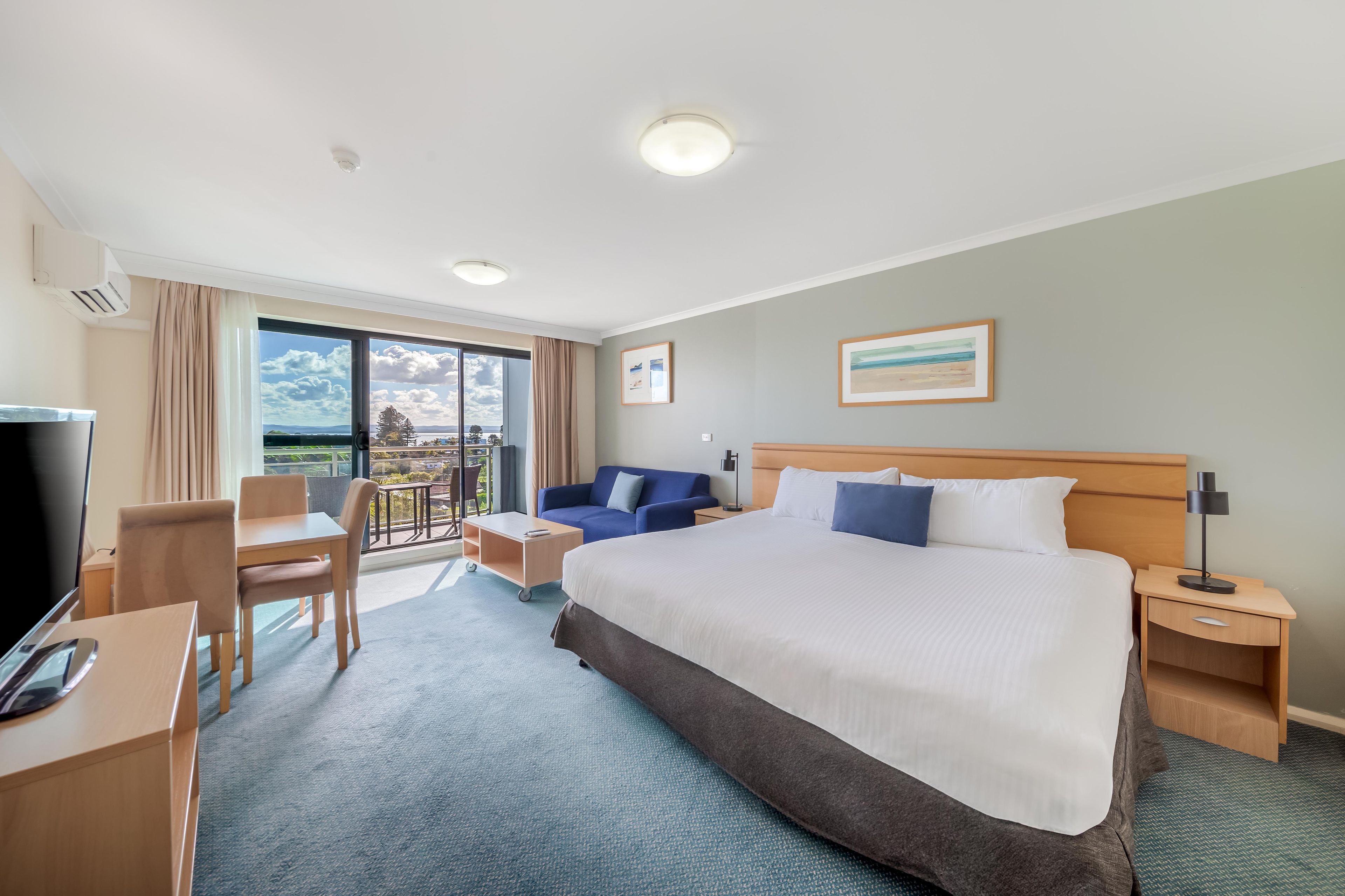 A hotel room with a bed, a desk, a chair, a TV, a balcony with a view, and a painting on the wall.