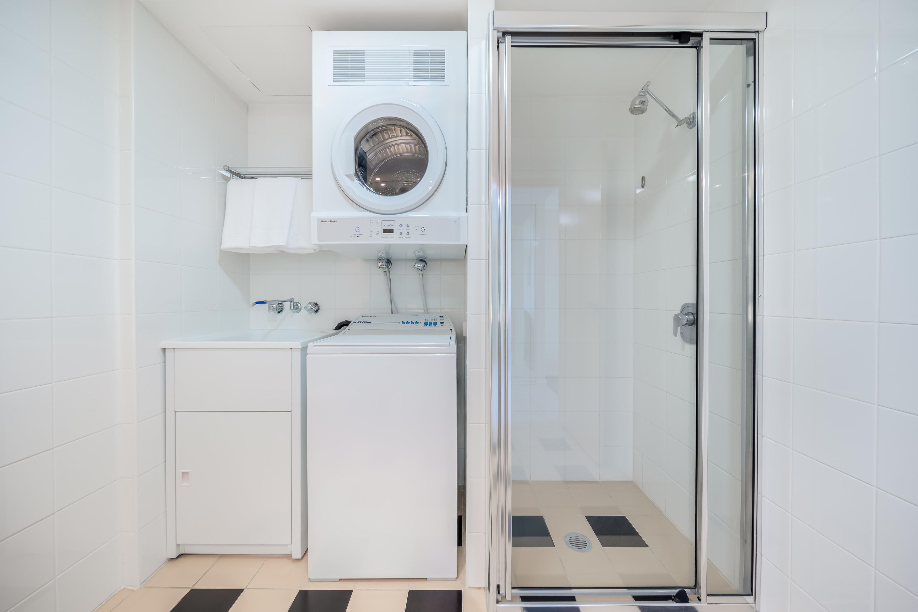 A compact laundry room with a front-loading washer and dryer, a glass-enclosed shower, and a small cabinet.