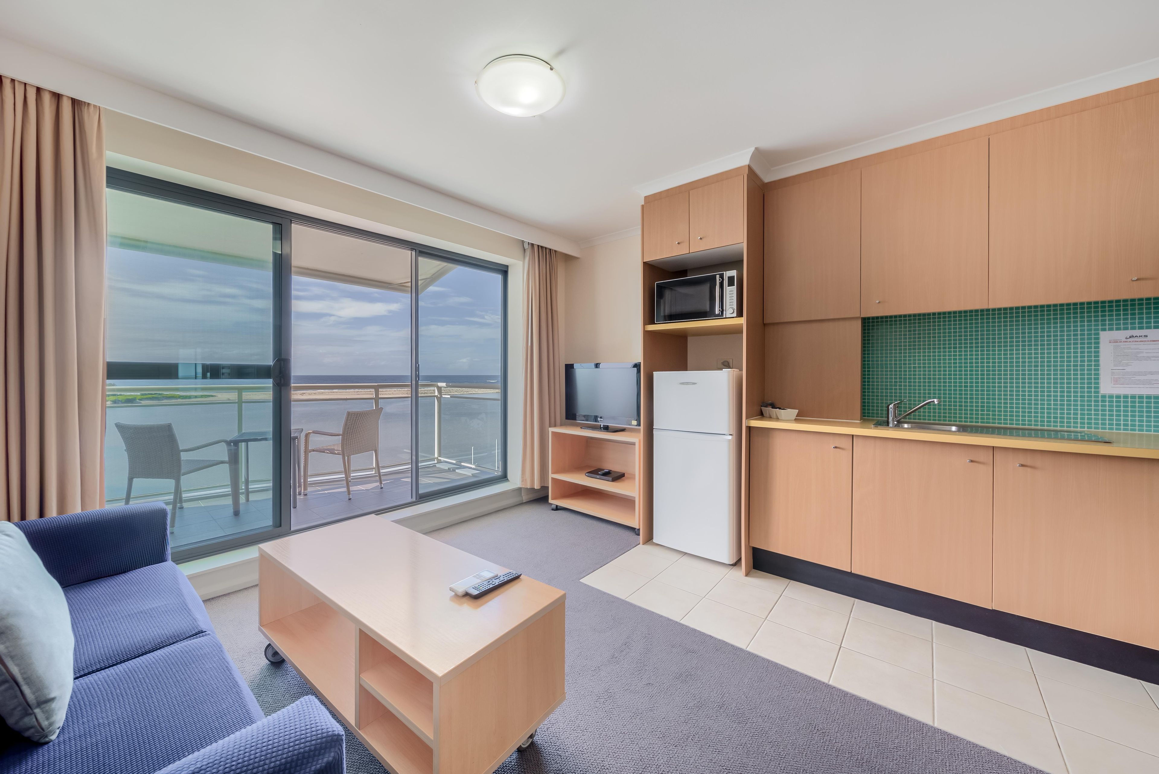 A modern hotel room with a balcony, a sofa, a small table, a television, a microwave, and a refrigerator.