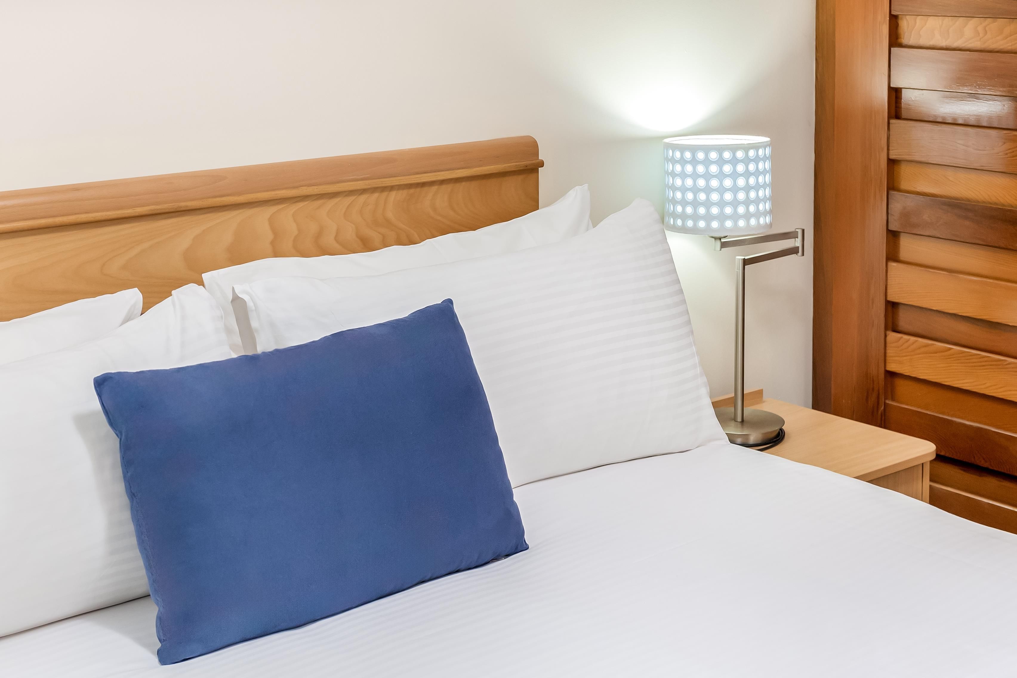 Una cama bien tendida con almohadas blancas y azules, un cabecero de madera y una lámpara de noche con pantalla estampada.
