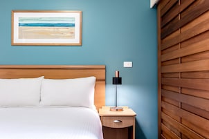 Una cama bien hecha con sábanas blancas, una mesita de noche con lámpara y un cuadro en la pared.