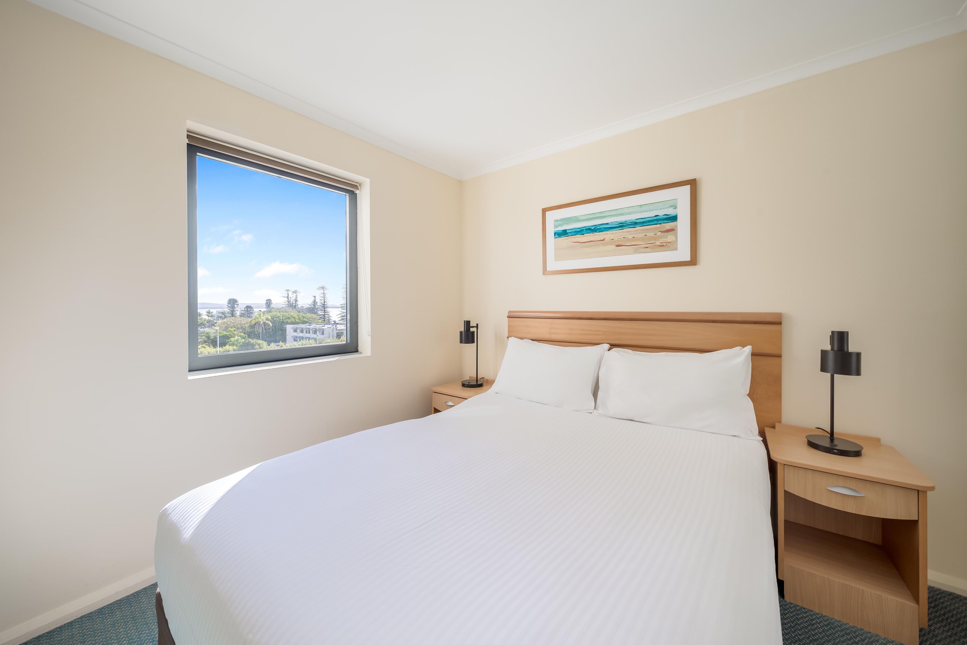 A hotel room with a large bed, a nightstand, a bedside table, a window with a view, and a framed picture on the wall.