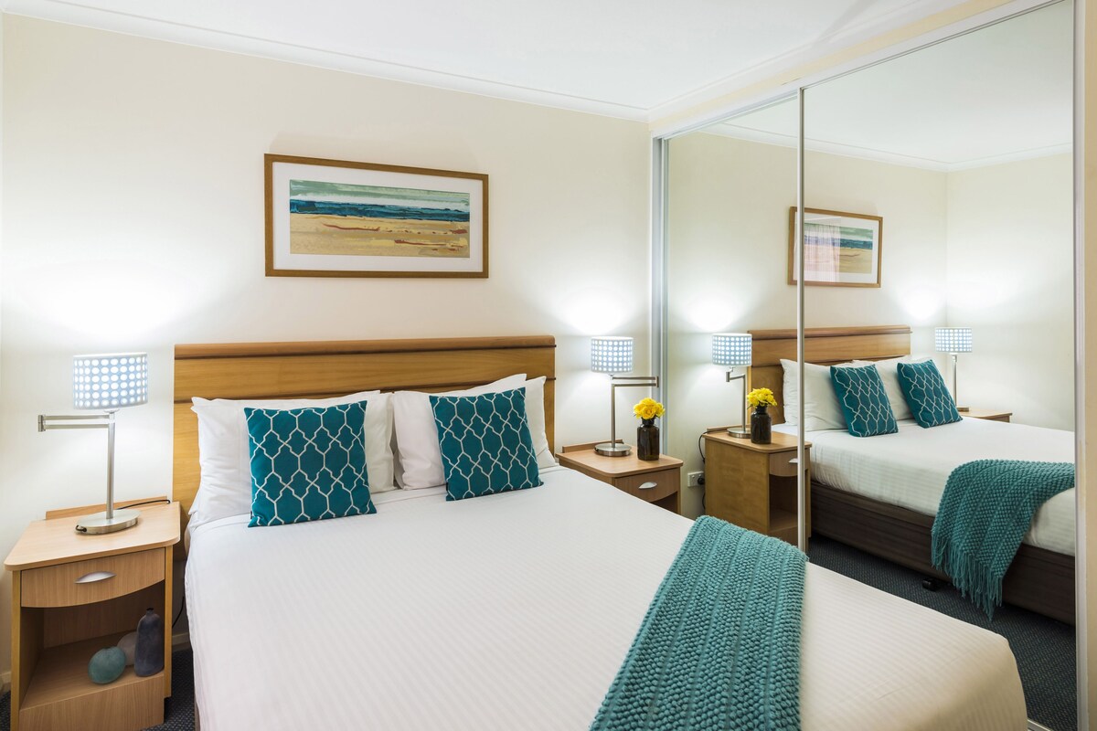 Un cuarto de hotel con una cama grande, dos mesitas de noche, un tocador, un espejo y un cuadro en la pared.