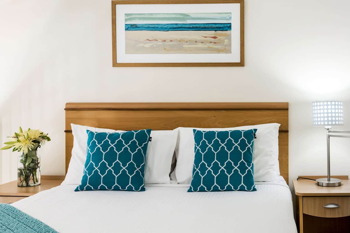 A neatly made bed with white and teal patterned pillows, a wooden headboard, a framed artwork of a beach scene, a bedside table with a vase of flowers, and a lamp on the nightstand.