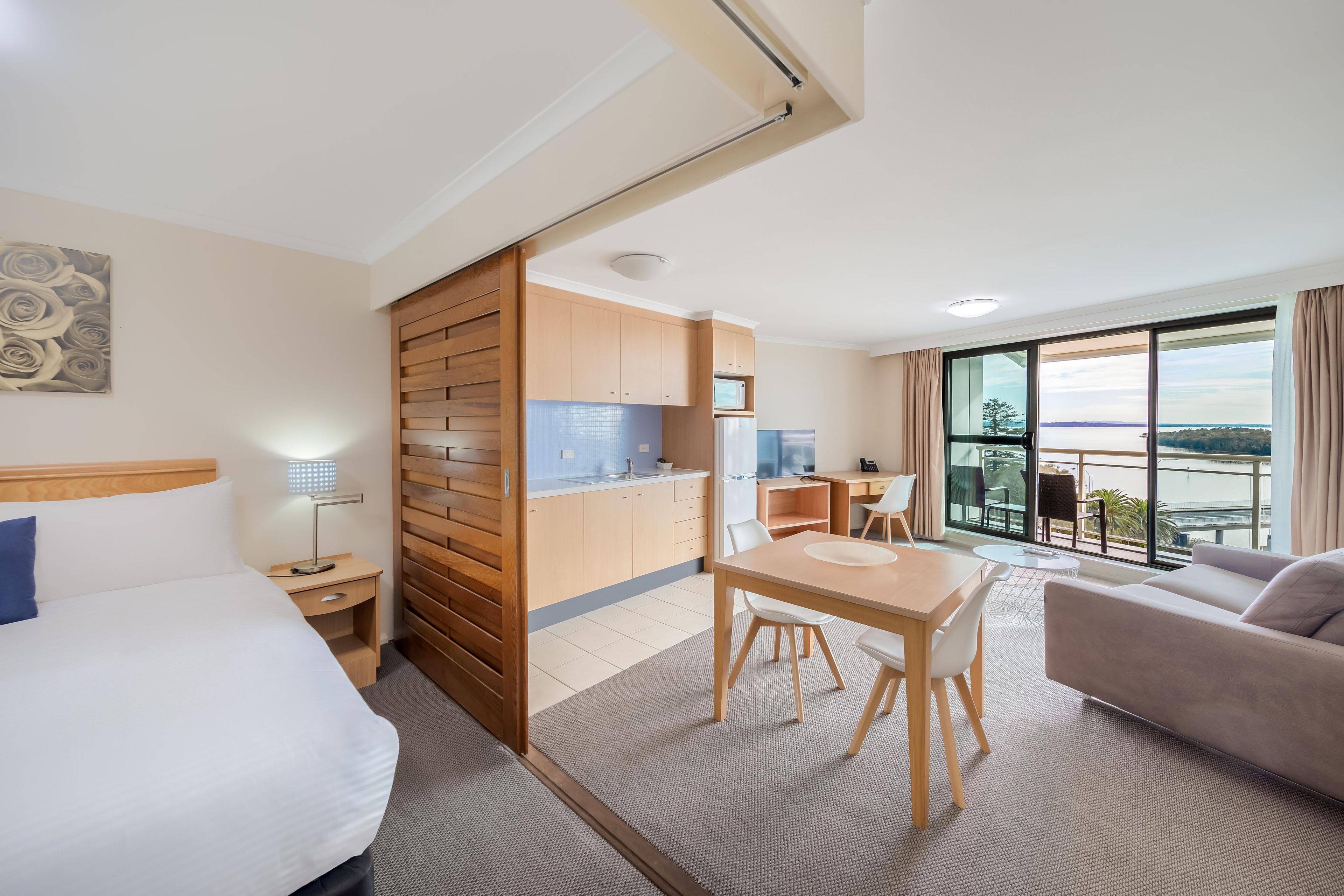 A hotel room with a bed, a desk, a dining area, and a balcony with a view.