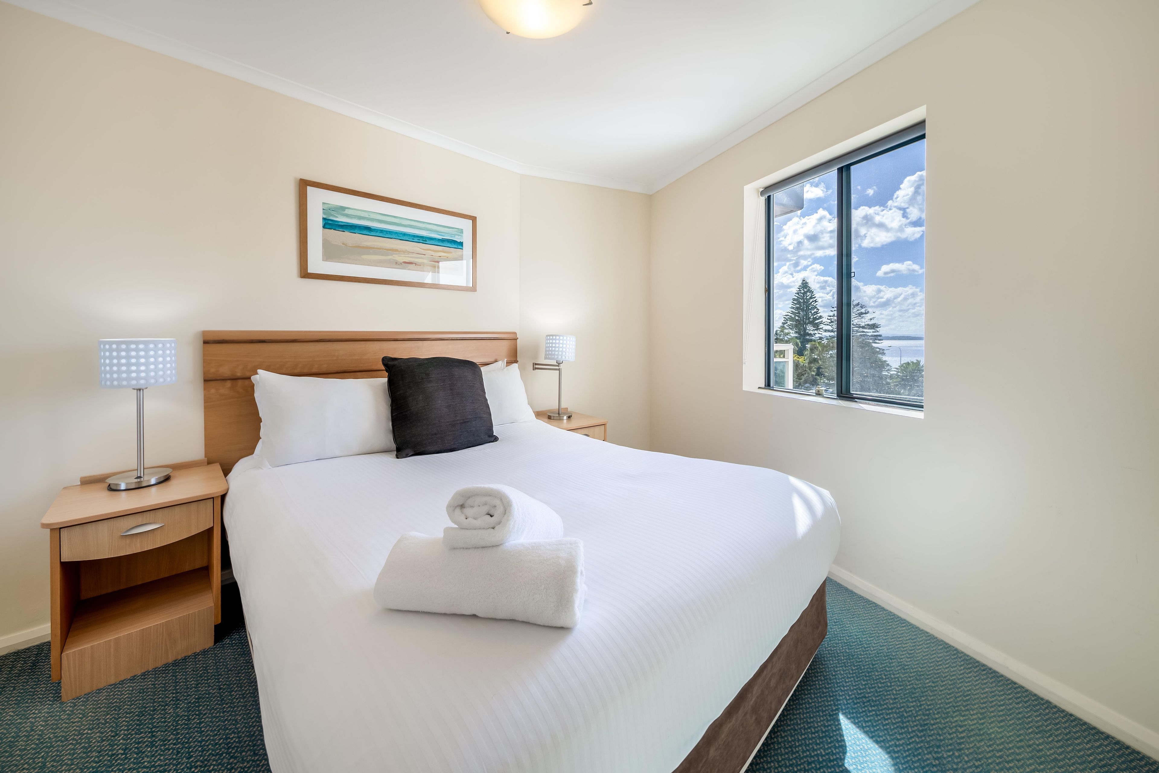 Habitación de hotel con una cama bien hecha, una mesita de noche con una lámpara, un cuadro en la pared y una ventana con vistas al cielo y nubes.