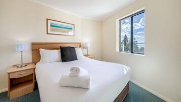 Habitación de hotel con una cama bien hecha, una mesita de noche con una lámpara, un cuadro en la pared y una ventana con vistas al cielo y nubes.
