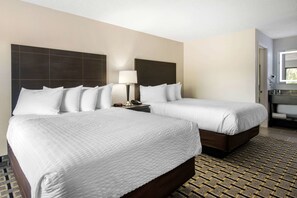 A hotel room with two beds, a nightstand with a lamp, a bathroom with a window, and a patterned floor. - Clarion Inn & Suites Kissimmee-Lake Buena Vista South (Kissimmee)
