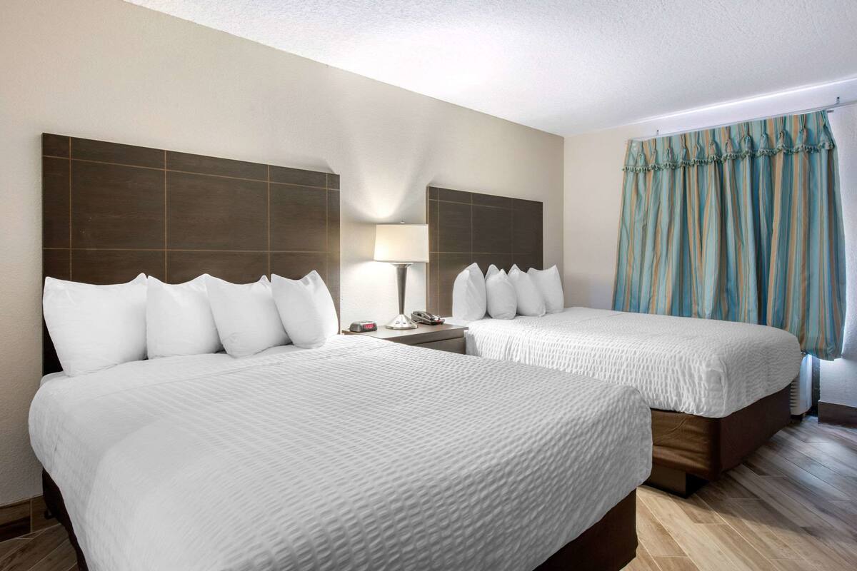 A hotel room with two beds, a nightstand, a lamp, and a window with curtains.