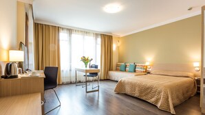A hotel room with a desk, two beds, a chair, a table with a vase of flowers, and a view of the outdoors. - Hotel Geneva (Sofia)