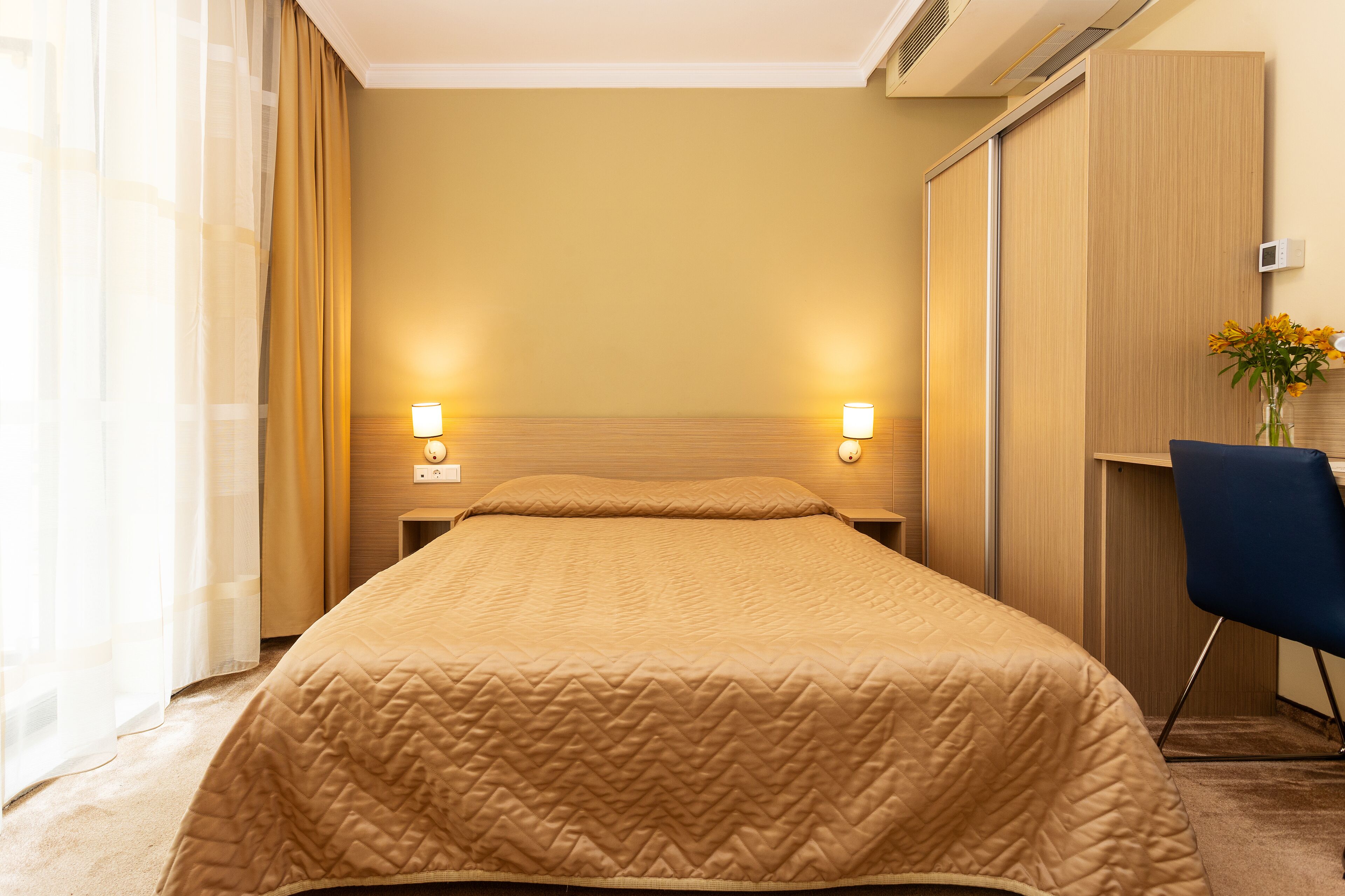 A hotel room with a large bed, two bedside lamps, a desk with a chair, and a vase with flowers.