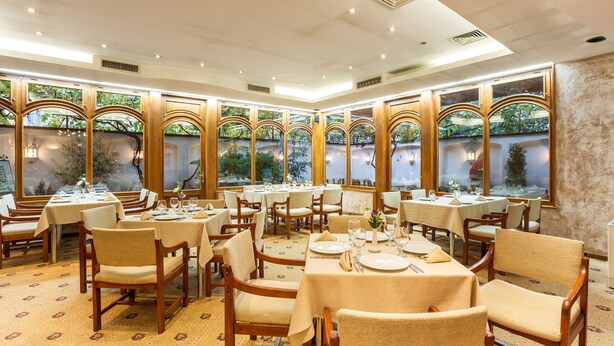 A spacious dining area with multiple tables and chairs, large windows with a view of greenery, and a well-lit interior. - Hotel Geneva (Sofia)