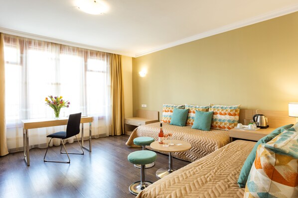 A hotel room with two beds, a desk, a chair, and a small table with a bottle and glasses.