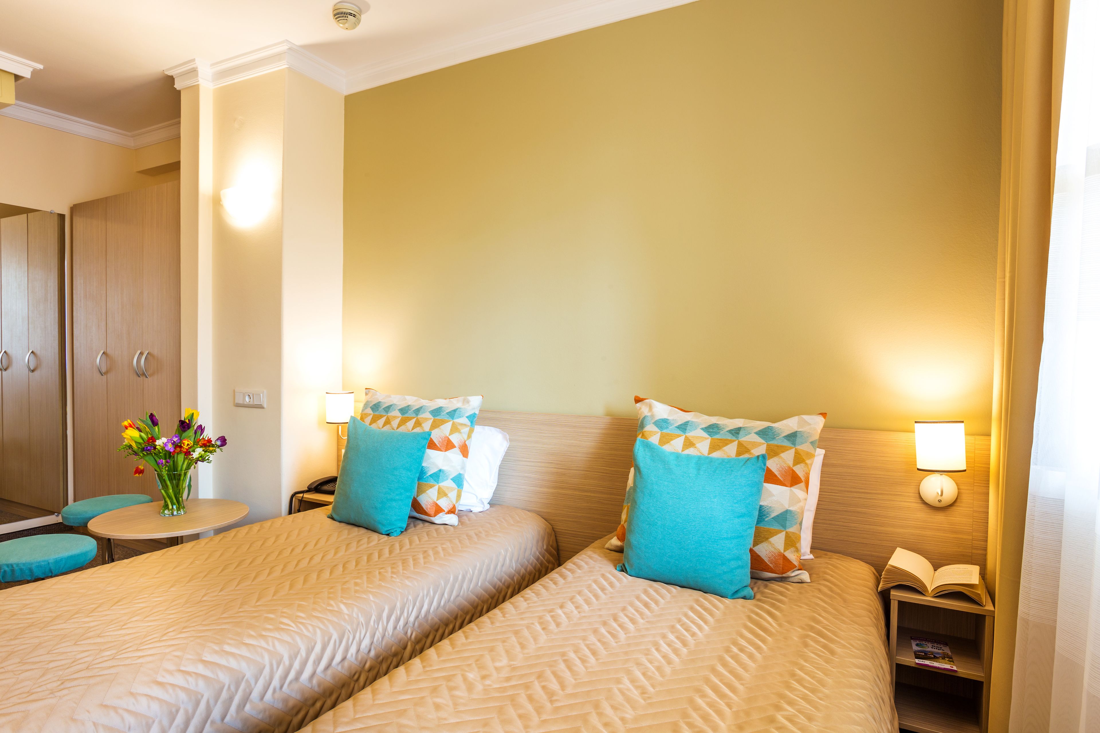 A hotel room with two beds, a small table with a vase of flowers, and a nightstand with a lamp and an open book.