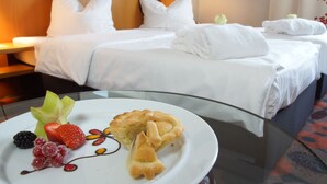 A hotel room with a bed, two bedside tables, and a glass table with a plate of fruit and a pastry.