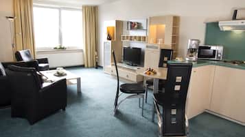 Habitación de hotel compacta con kitchenette, zona de comedor con mesa y sillas, sofá y ventana grande con cortinas.