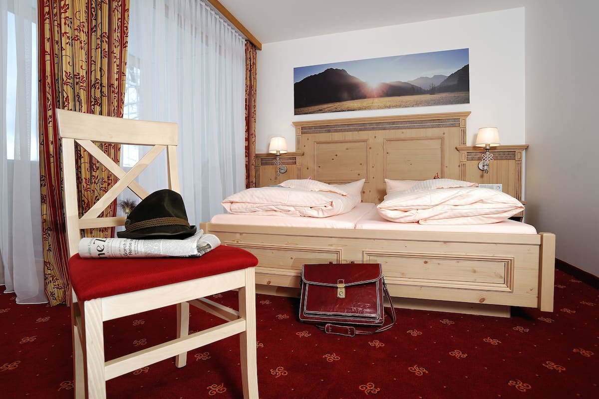 A hotel room with a wooden bed, a red chair, a handbag, and a painting of a mountain landscape on the wall.