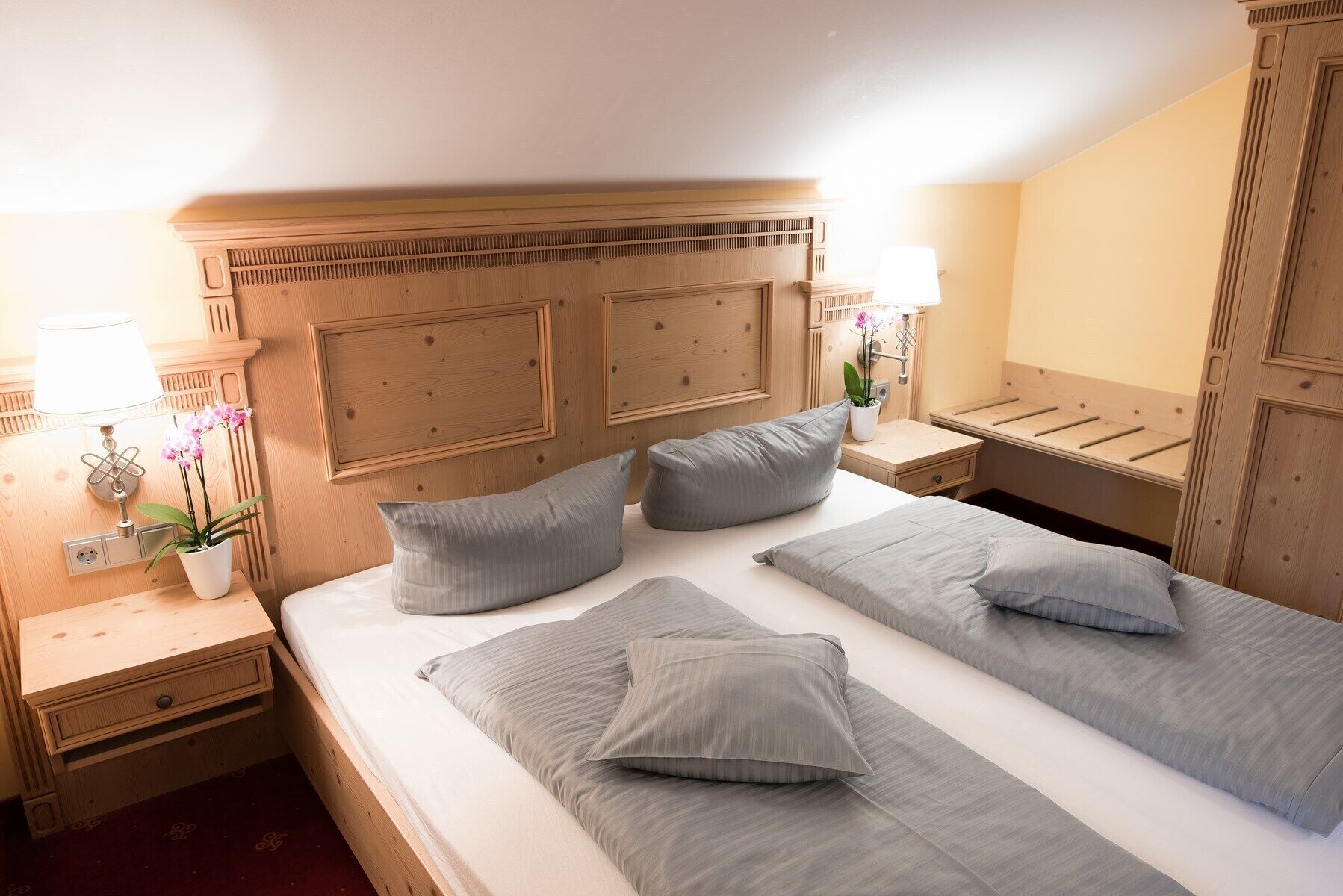A hotel room with a wooden headboard, two beds with grey bedding, a nightstand with a lamp, a small table with a potted orchid, and a wooden wardrobe.