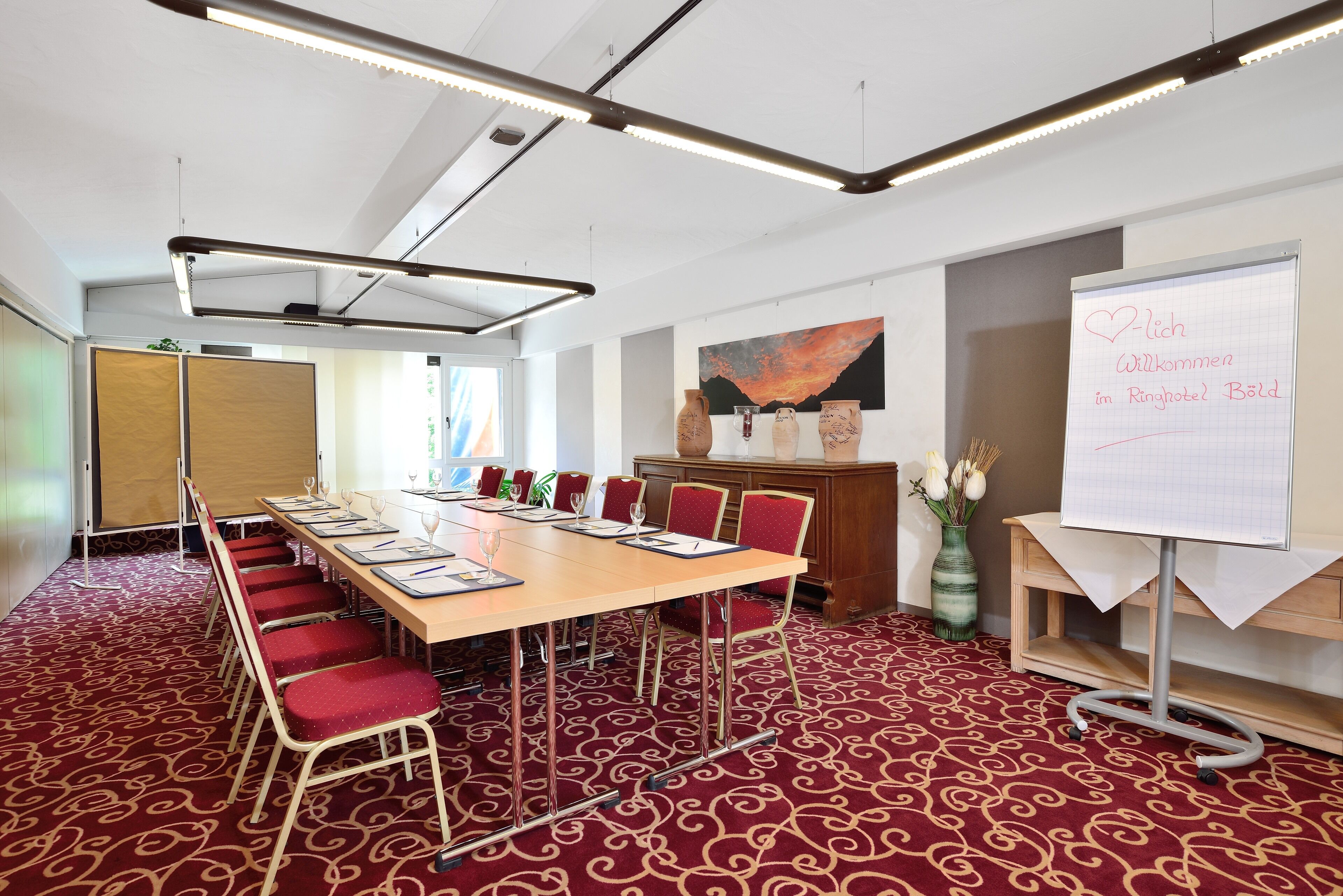 A conference room with a long table, red chairs, a whiteboard, and a decorative vase.