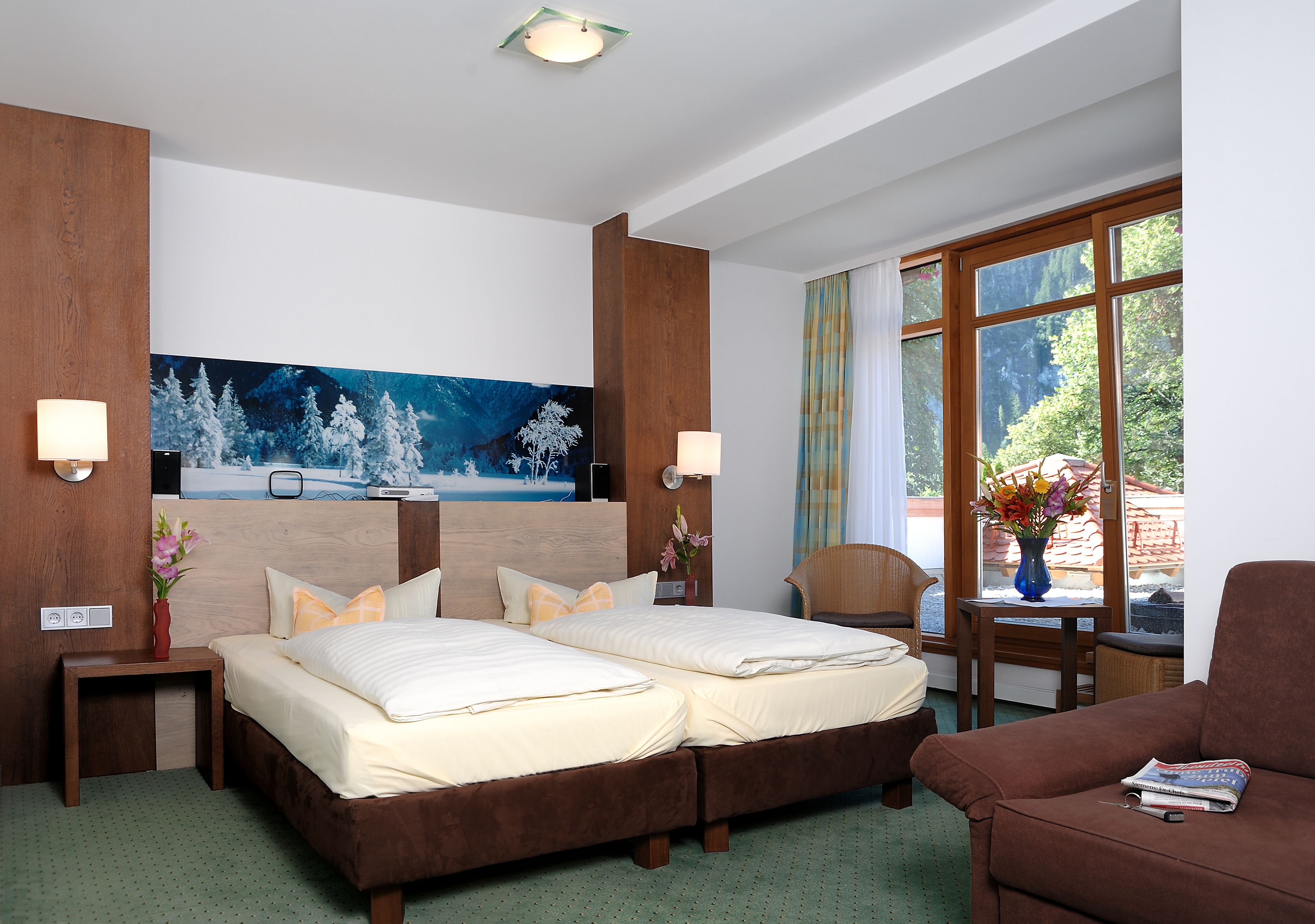 Ein Hotelzimmer mit einem großen Bett, zwei Nachttischen, einem Schrank, einem Holz-Kopfteil, einem Fenster mit Vorhängen, einem Sessel und einem Bild an der Wand.
