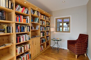 A room with wooden bookshelves filled with books, a red plaid armchair, a small table with a lamp, and a window with a view. - Landhotel Böld and Restaurant Uhrmacher Oberammergau (Oberammergau)