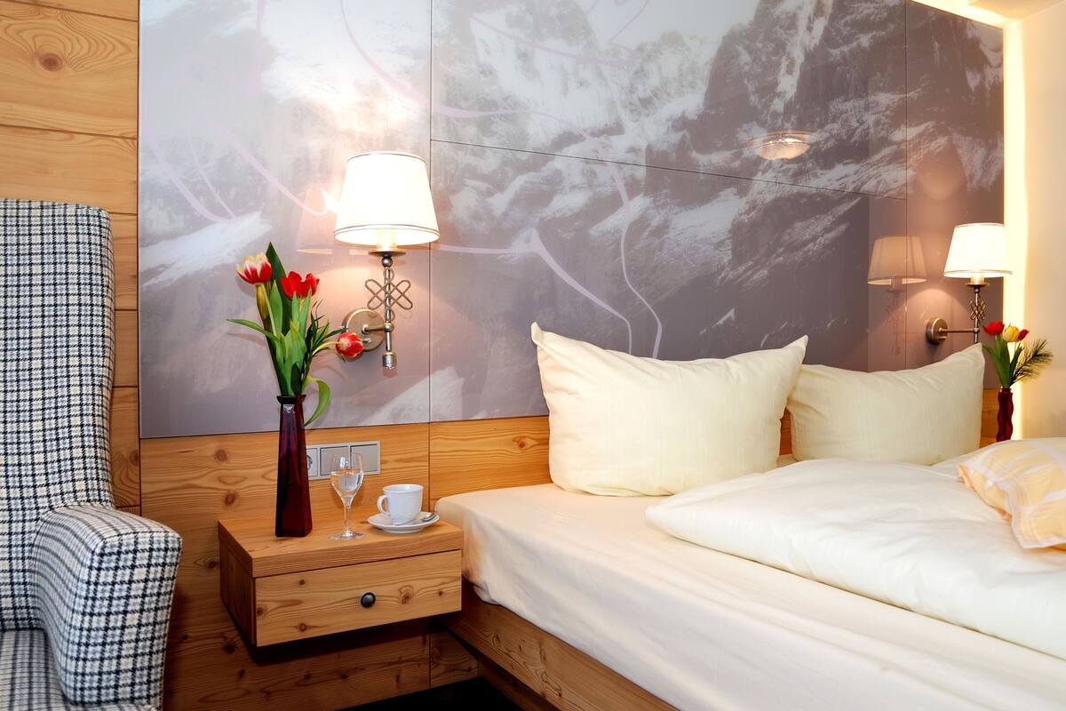 A hotel room with a bed, a nightstand, a lamp, a vase with flowers, and a chair.