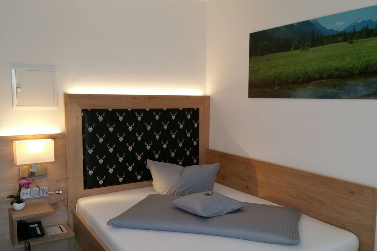 A bedroom with a bed, a nightstand with a lamp, a small table with a vase of flowers, and a painting of a landscape on the wall.