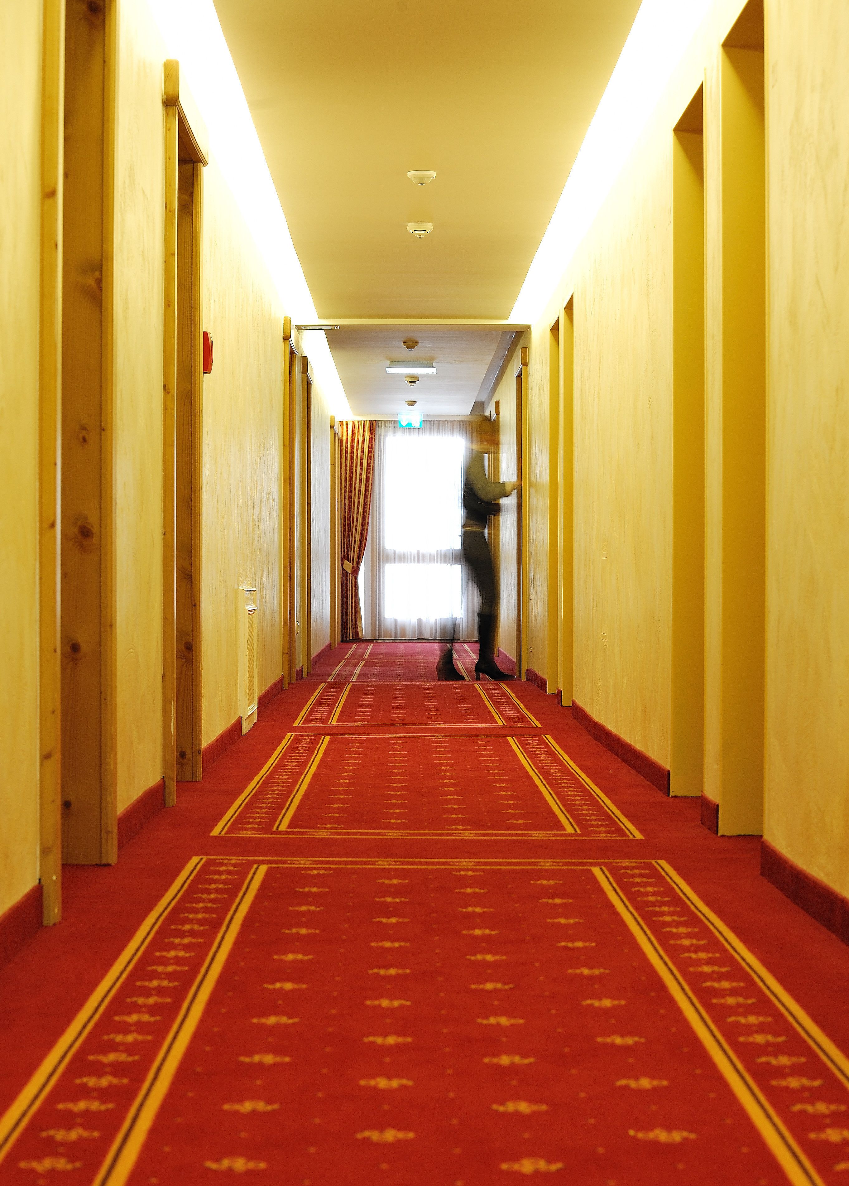 A hotel hallway with a red carpet, wooden walls, and a person walking towards the end.