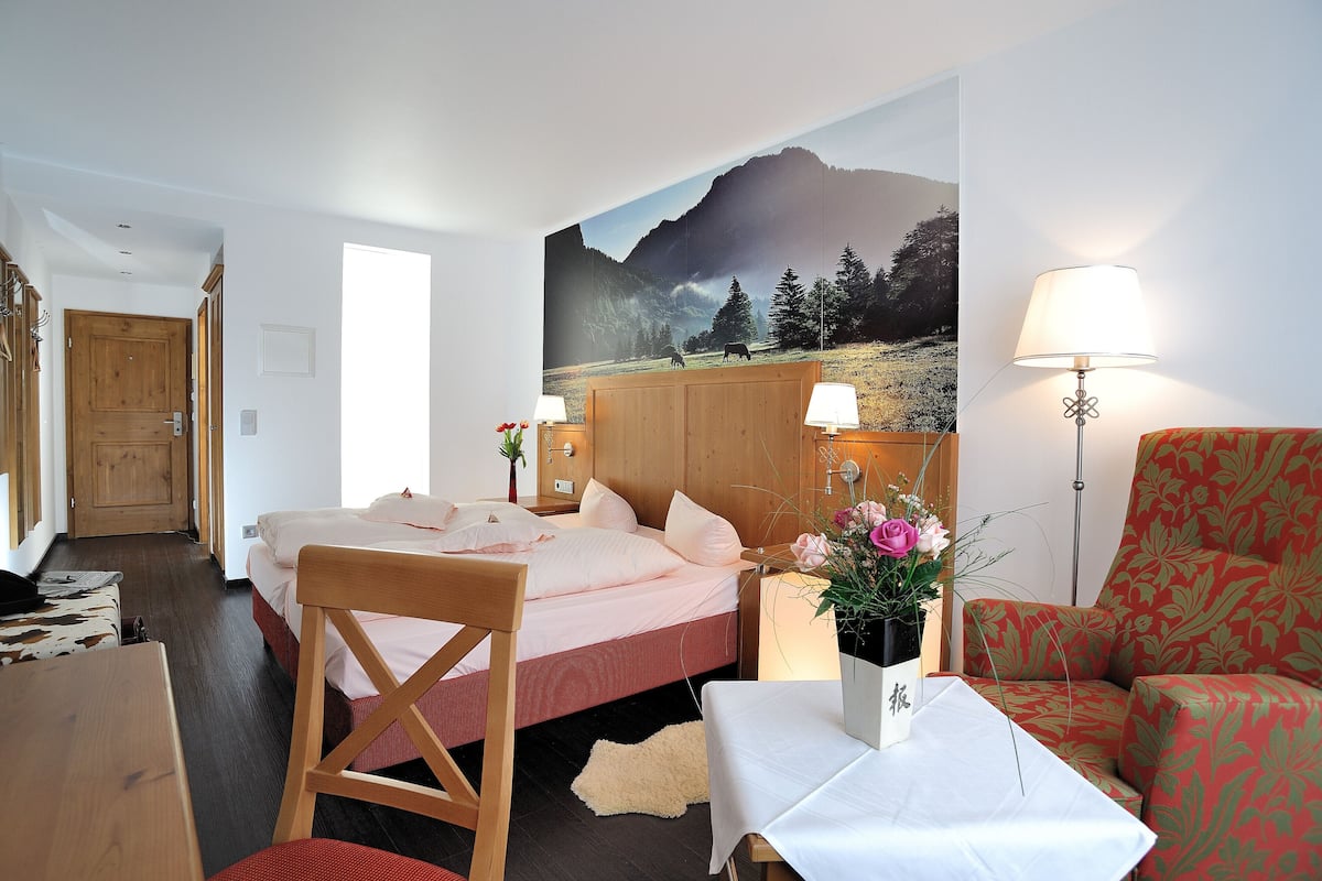 A hotel room with a bed, a chair, a table, a lamp, and a painting of a mountain landscape.