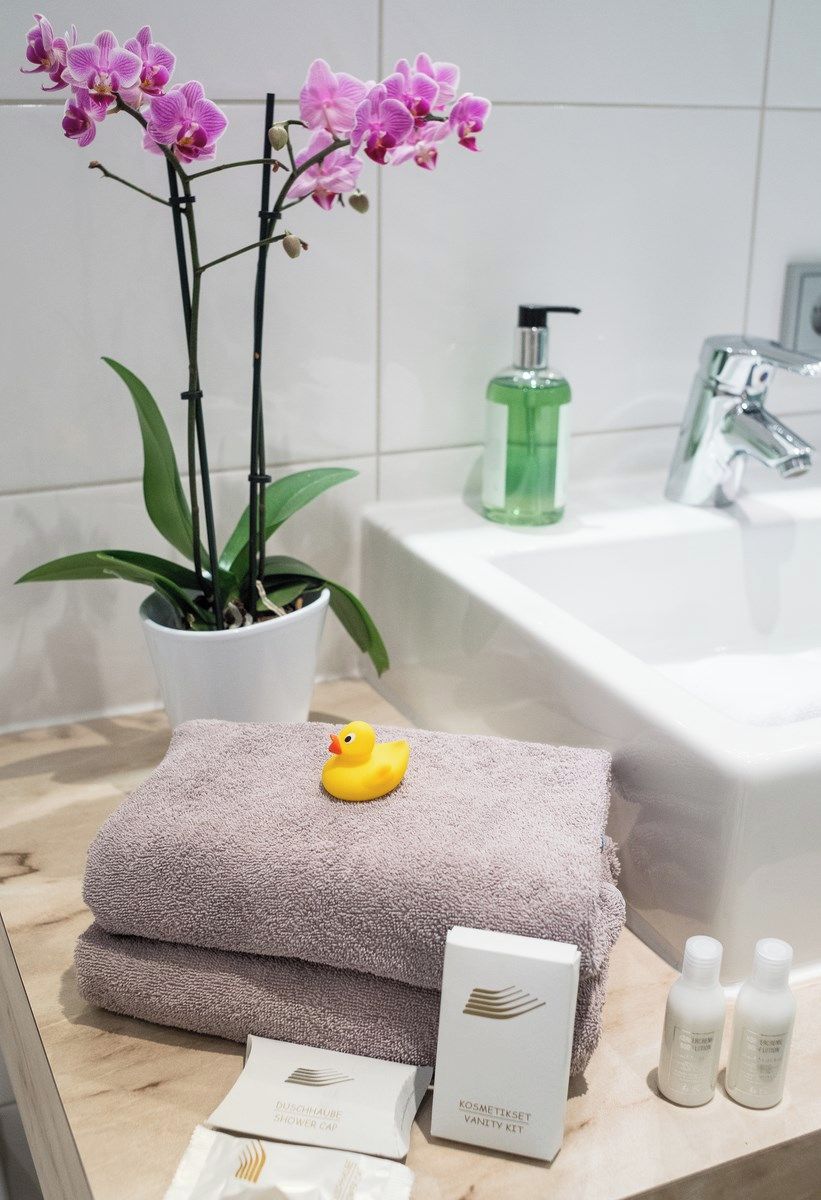 Ein Badezimmer mit einem weißen Waschbecken, einer grünen Flasche Flüssigseife, einer rosa Orchideenpflanze, einem gelben Gummibadenten, einem gefalteten Handtuch und einer Schminktasche.