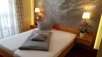 En pænt redt seng med puder og et foldet håndklæde, to sengeborde med lamper, et natbord med en plante, og en væg med marmorlignende struktur.