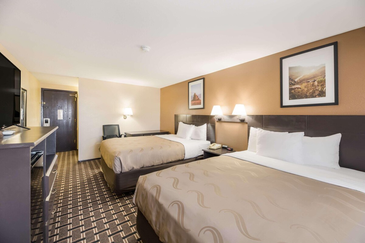 Quarto de hotel com duas camas, uma escrivaninha, uma cadeira, uma televisão e um quadro na parede.