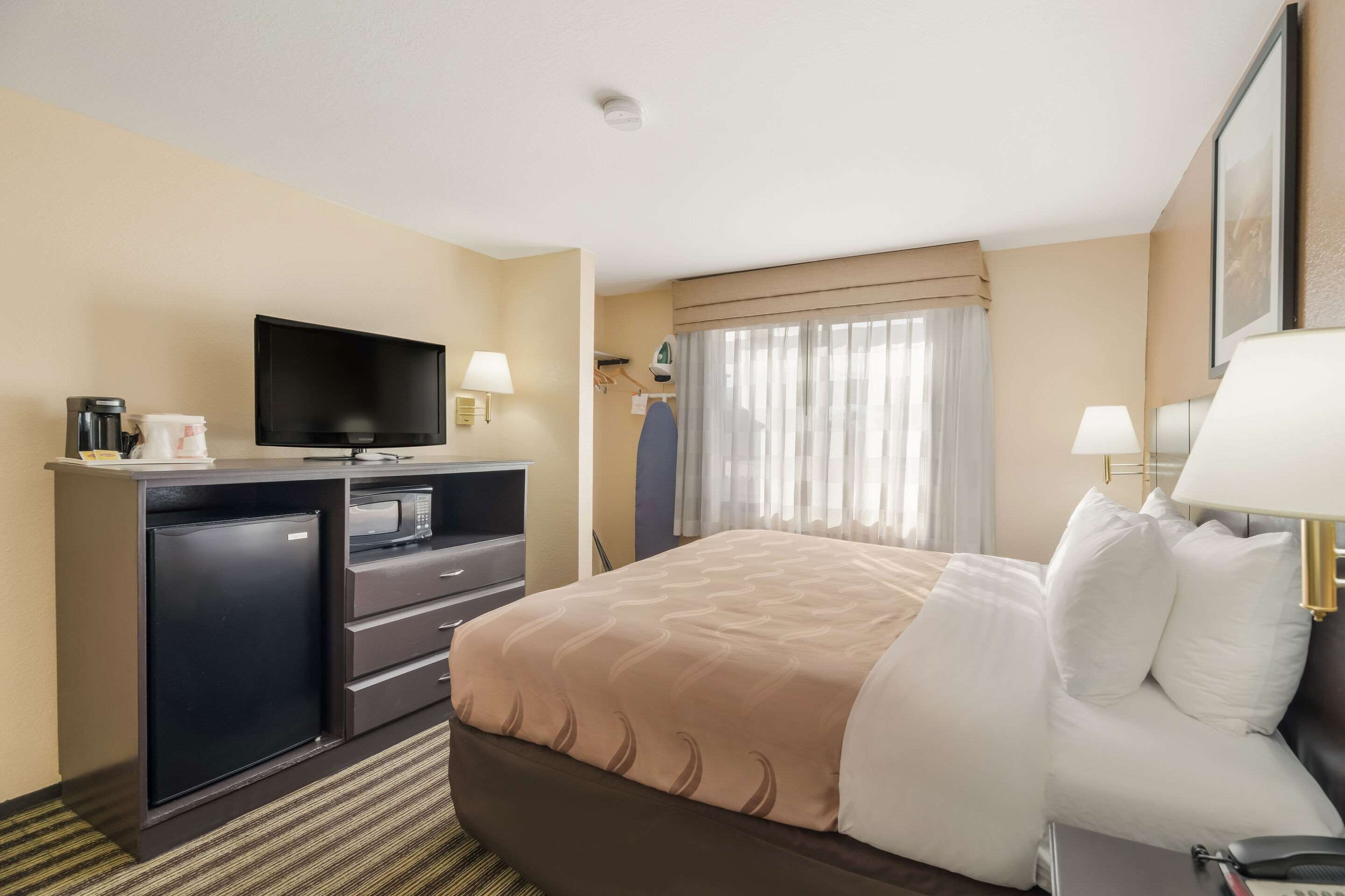 A hotel room with a bed, a TV, a desk with a computer, a coffee maker, and a refrigerator.