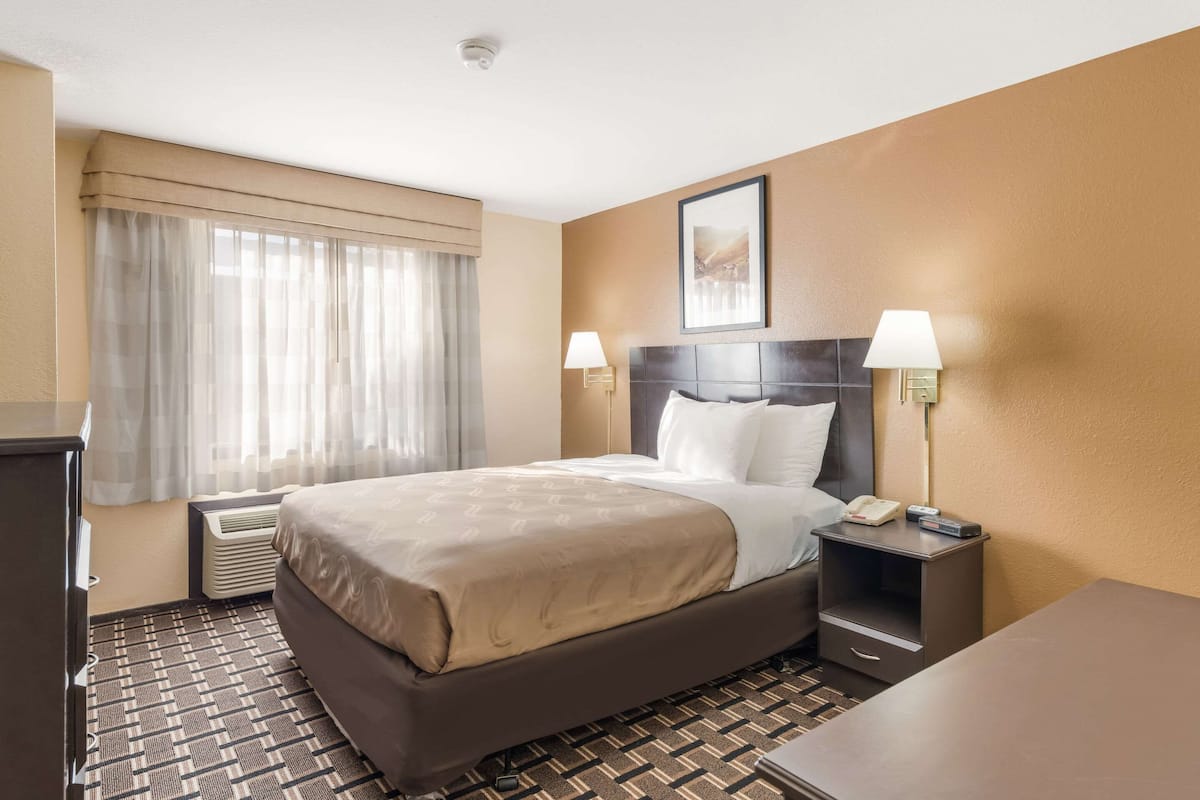 Quarto de hotel com uma cama, duas luminárias de cabeceira, uma mesa de apoio, uma escrivaninha e uma janela com cortinas.