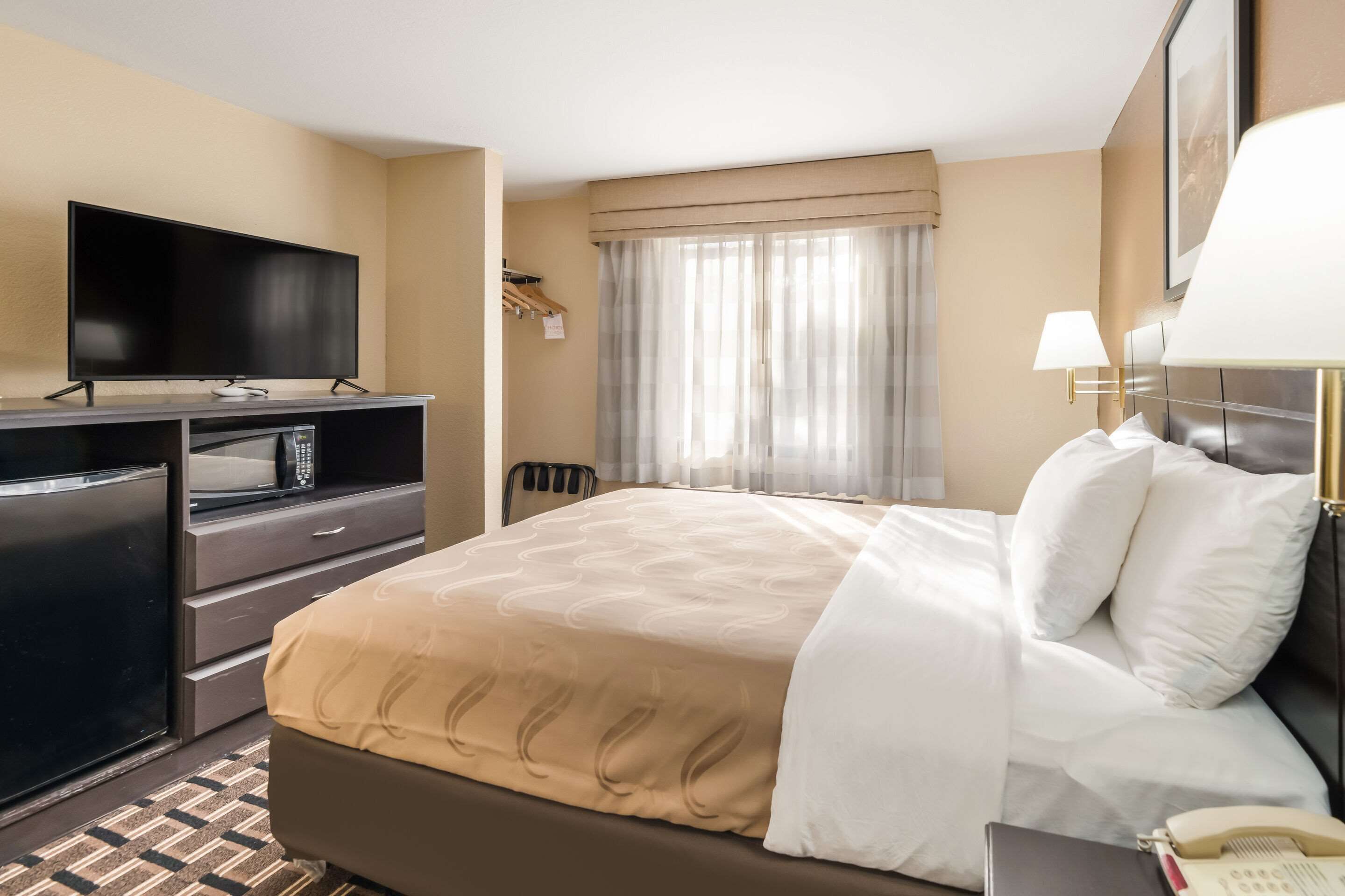 Habitación de hotel con cama, televisión, escritorio con ordenador, teléfono y ventana con cortinas.