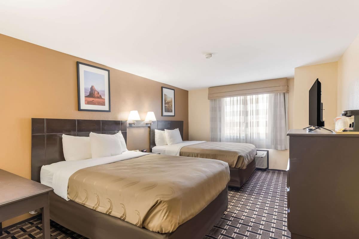 Quarto de hotel com duas camas, uma escrivaninha, televisão e janela com cortinas.