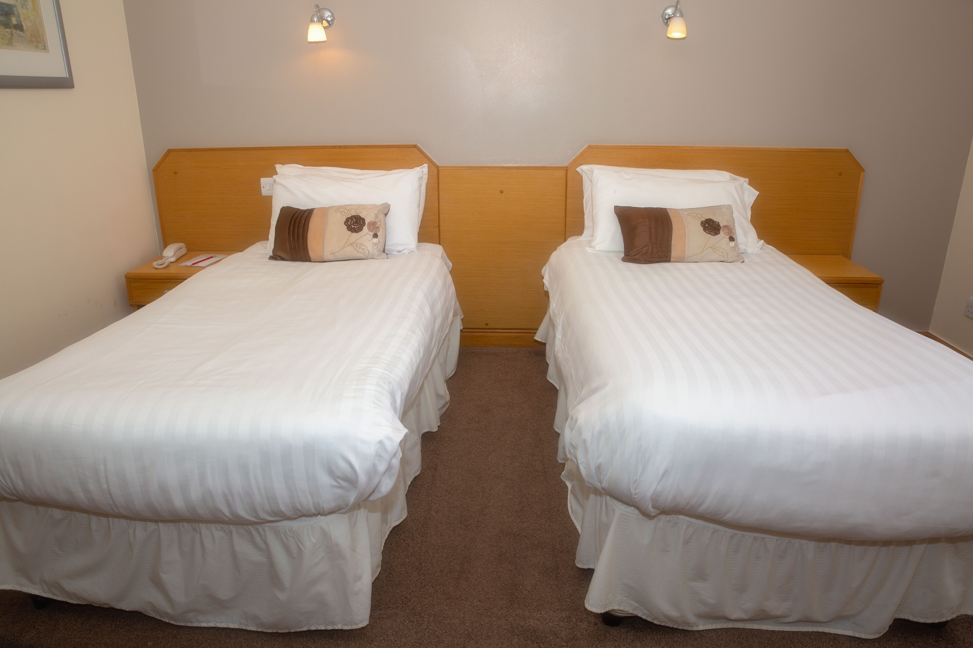 A hotel room with two single beds, each with a wooden headboard, white bedding, and a nightstand with a telephone and a book.