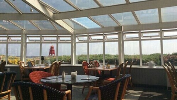 A sunlit dining area with glass walls, wicker chairs, and a view of a red structure in the distance. - Little Haven Hotel On The Beach (South Shields)