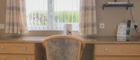 Una habitación con escritorio de madera, dos cajones, una silla, ventana con cortinas, televisor de pantalla plana montado en la pared y toma de corriente.