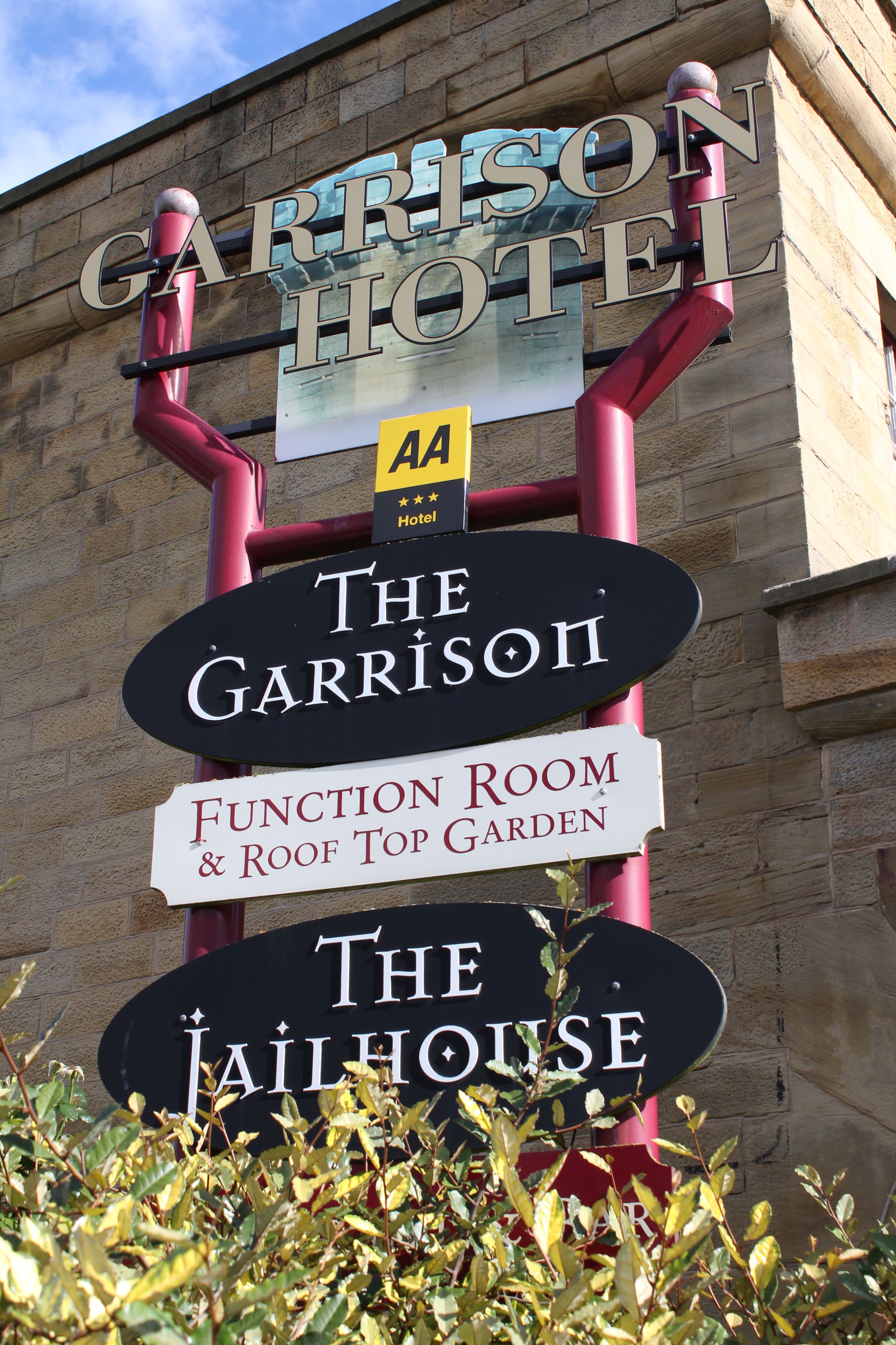 Een hotelschild met verschillende namen, waaronder 'Garrison Hotel', 'The Garrison', 'Function Room & Roof Top Garden' en 'The Jailhouse'.