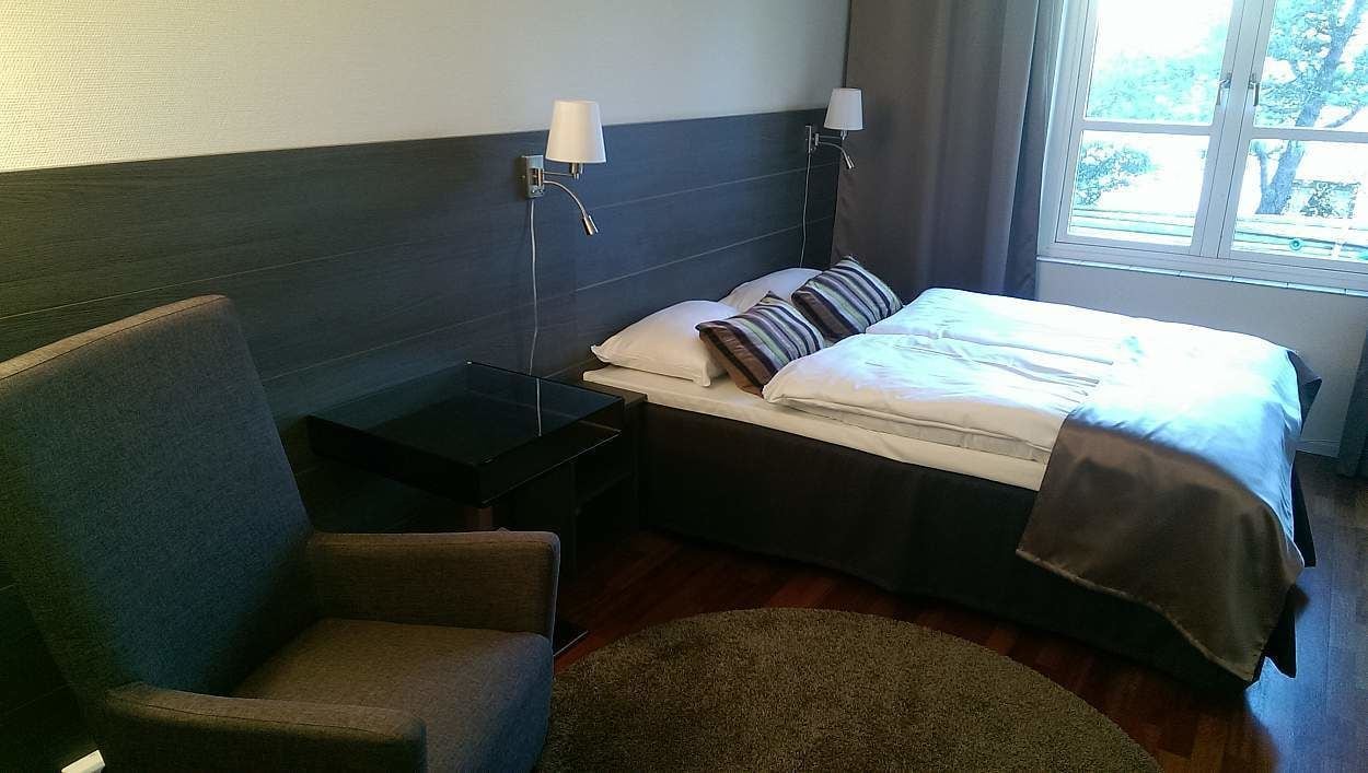 Una habitación de hotel con una cama, una silla, una mesita de noche y una ventana.