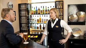Uma área de bar com um barman e um cliente. Há prateleiras com vários copos e garrafas atrás do balcão.